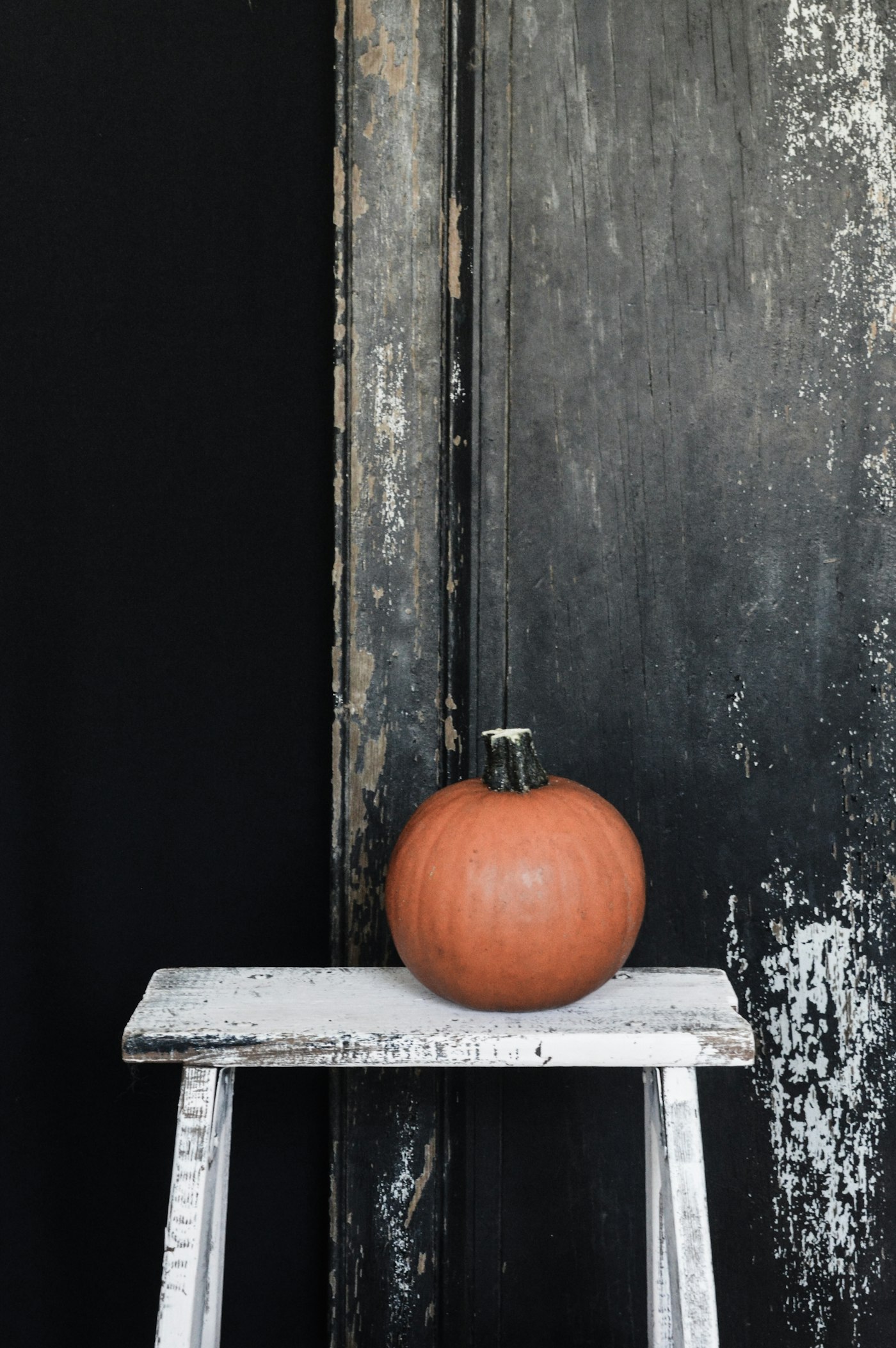 One orange pumpkin on a chippy white stool against a dark weathered wall