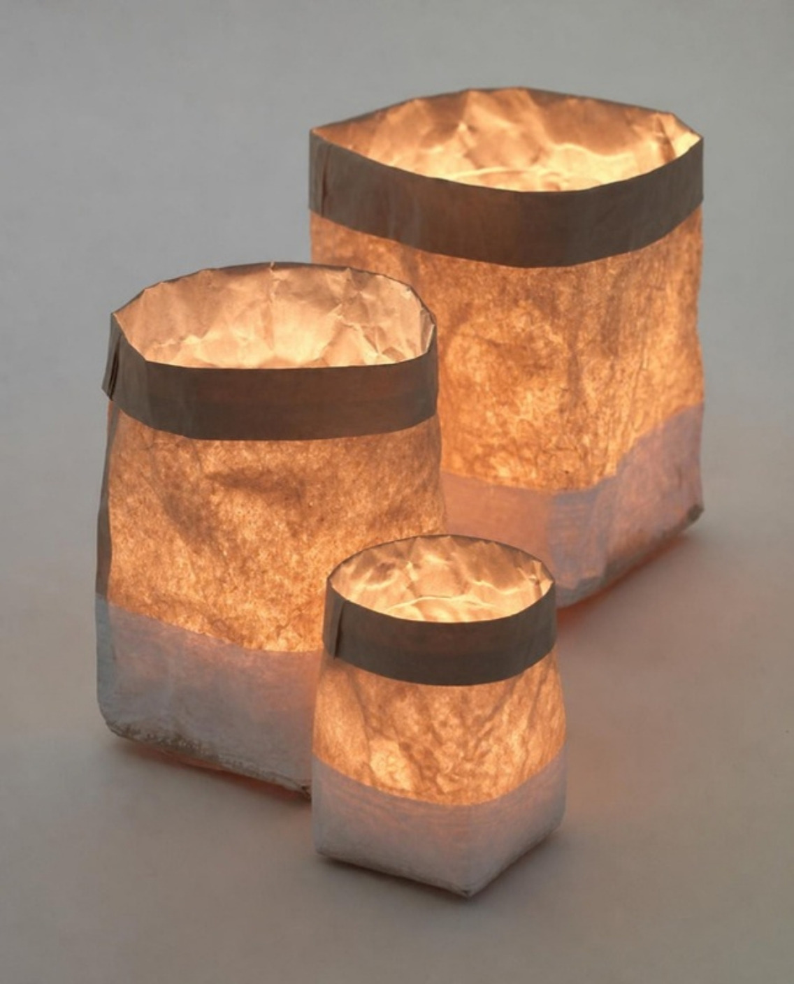 Three crinkled paper bag lanterns with gray folded tops, glowing warm from inside on a soft gray background