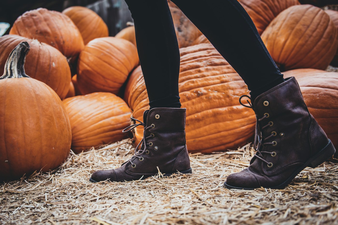 Someone in brown lace-up boots and black jeans walking on straw past a pile of big orange pumpkins