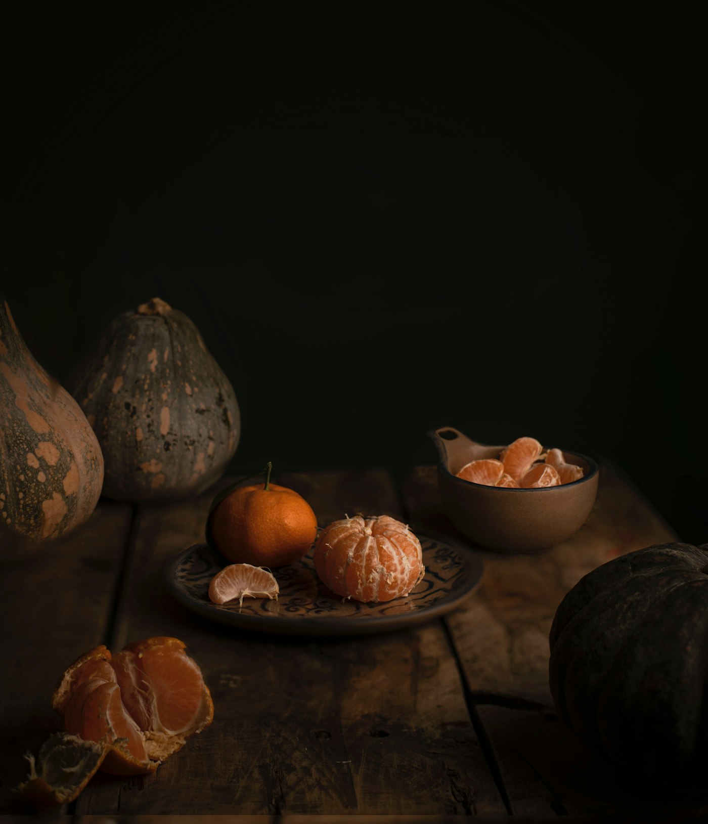 Old speckled squash, a handmade plate and bowl with peeled tangerines on a worn wooden table in low light