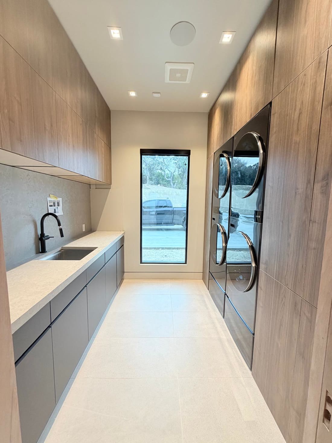 The scullery in pale timber and gray, with stacked machines and a window to the drive