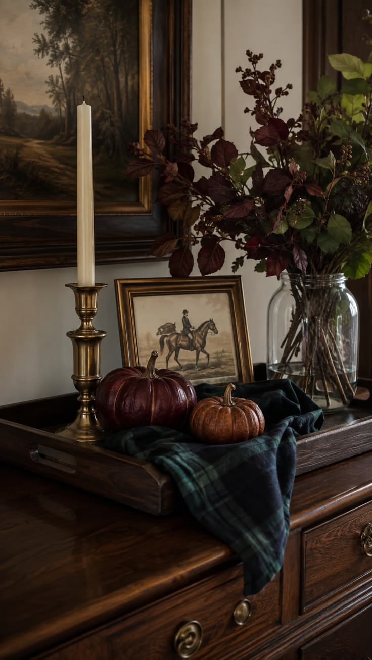 An old wood tray on an antique dresser with a brass candlestick, a horse print in a gilt frame, a plum and a rust pumpkin, a plaid throw and burgundy leaves in a glass vase