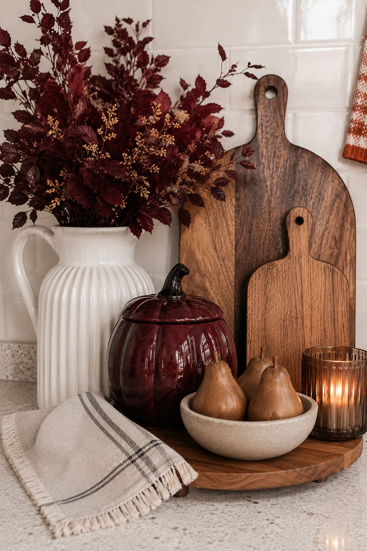A kitchen counter corner with deep burgundy leaves in a ribbed white pitcher, wood cutting boards, a burgundy pumpkin jar, pears in a bowl, a candle and a striped linen towel