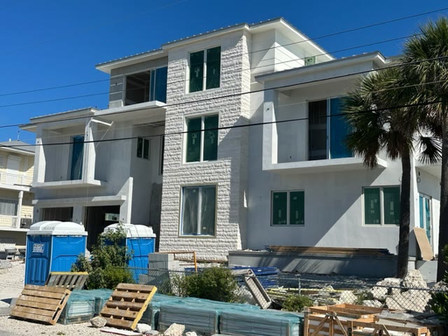 The house under construction from the side, stepped balconies and the stone center, materials stacked in the foreground